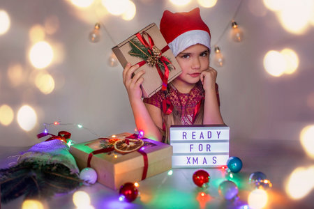 Cute small girl in red Santa hat waiting for miracle before Christmas season, festive illumination, garland and packed gifts, she is ready for celebration, indoor portrait over white backgroundの写真素材