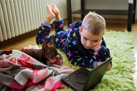 7-8 year old boy lying on a rug next to a battery while watching a movie on a compact DVD player. Chihuahua dog also attentively watches the movie with himの写真素材
