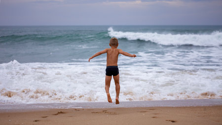 A boy of 7-8 years old sits on the ocean shore with his arms open towards the wind and waves. Storm on the ocean in summer. The concept of freedom, courage and challenge. Place for textの写真素材