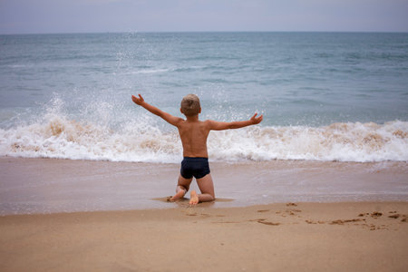 A boy of 7-8 years old sits on the ocean shore with his arms open towards the wind and waves. Storm on the ocean in summer. The concept of freedom, courage and challenge. Place for textの写真素材