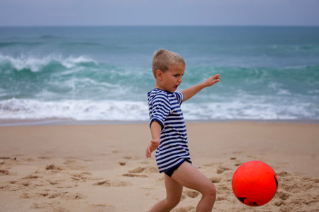 A small football fan barefoot plays football just on the band of ocean. Football everywhere. Sport, soccer, fandom concept. Storm outside and storm of emotions insideの写真素材