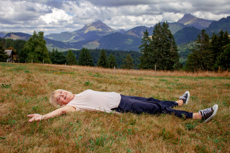 Elderly woman lays among the mountains with outstretched arms, overwhelmed with emotions. Beautiful landscape. Active aging, inner peace and tranquilityの写真素材