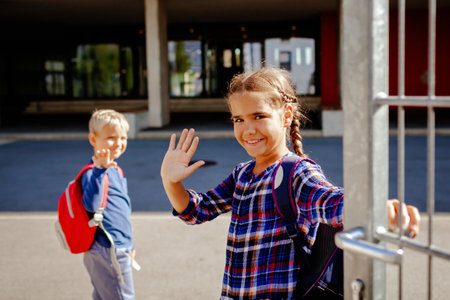 Happy siblings with backpacks, primary school kids, waving their hands near the school at first day, study and education, reunion after vacations, lifestyle. Focus on girl. Back to schoolの写真素材
