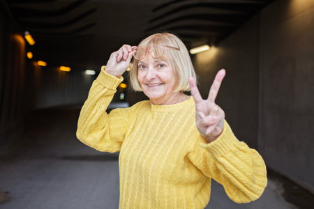 Portrait of woman over 65 with eyeglasses in yellow autumn sweater shows victory gesture. She is cheerful and positive. Beautiful aging and an optimistic outlook on life.の写真素材