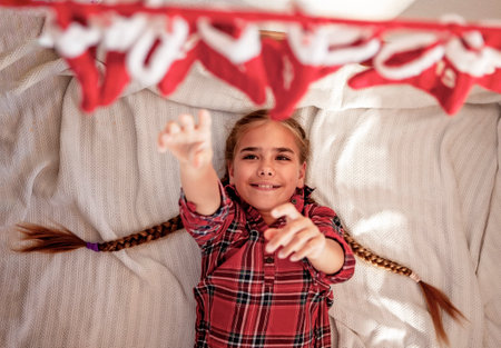 A cheerful girl with braided hair in plaid shirt lies underneath a hanging Advent calendar, reaching up eagerly to retrieve the days gift from a Santa Claus stocking. Festive excitement. Top viewの写真素材