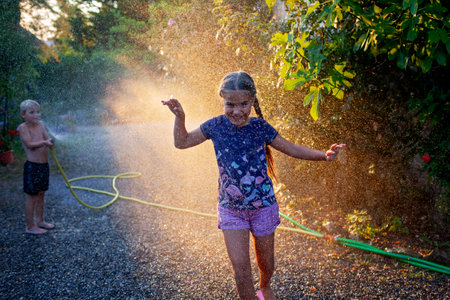 Joyful kids play with water hose in summer light, splashing and laughing in a backyard, creating memories in the golden hour sunlight.の写真素材