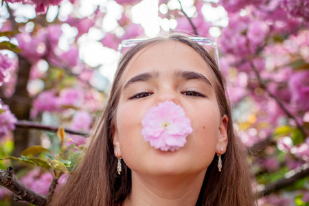 A teenage girl playfully holds a cherry blossom near her nose against a backdrop of blooming trees.の写真素材