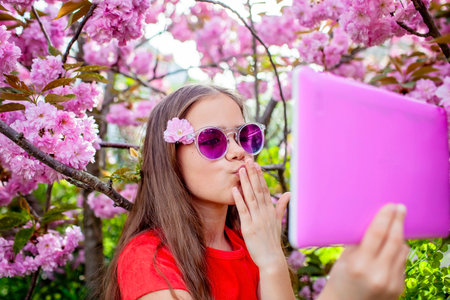 A girl sends an air kiss to a relative during a video call, surrounded by cherry blossomsの写真素材