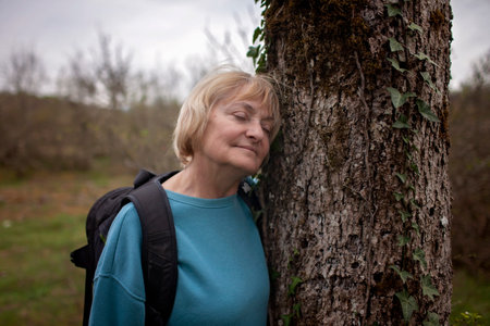 A senior woman with a serene expression rests against an old tree trunk, eyes closed, immersed in the tranquility of nature during a hiking break.の写真素材