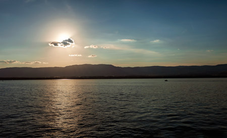 The sun sets behind a cloud over a calm lake with mountains in the distance, casting a tranquil scene.の写真素材