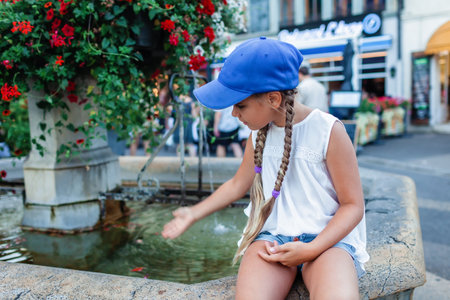 A young girl in a blue cap sits by a stone fountain, gently touching the water. Vibrant flowers cascade from the fountain, adding color to an urban settingの写真素材