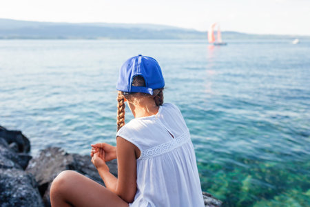 A young girl in a blue baseball cap and white dress enjoys a serene moment by the lake, watching sailboats glide across the water.の写真素材
