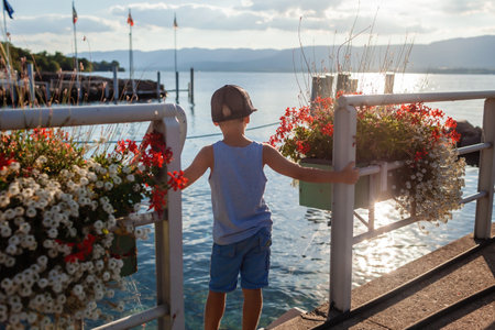 A young boy stands on a pier in a small village on Lake Leman, surrounded by ornate flower boxes, enjoying the tranquil scenery and the gentle touch of the sunset on the water.の写真素材