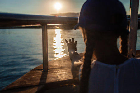 Child in a blue cap at sunset on a boat, touching sunlight reflection on the water, evoking a sense of wonder and the beauty of nature.の写真素材