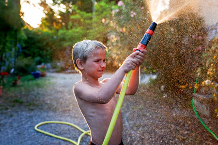 A young boy with joyful laughter sprays water from a garden hose, sparkling droplets flying in the warm sunlight, a quintessential summer moment captured.の写真素材