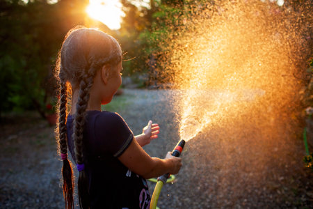 A joyful girl plays with a garden hose, spraying water in the golden light of the setting sun, creating a misty spray against a background of trees.の写真素材