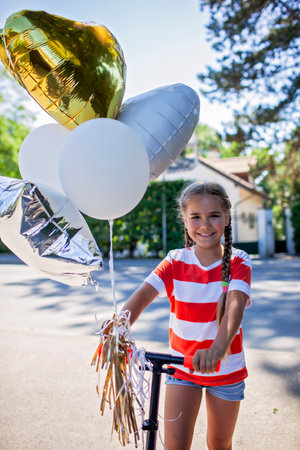 Young child riding a scooter outdoors during sunset. Her birthday balloons create a lively accent. This photo reflects happiness, motion, and summer freedom in a natural setting.の写真素材