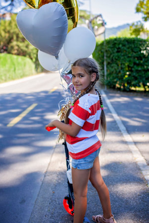 Young child riding a scooter outdoors during sunset. Her birthday balloons create a lively accent. This photo reflects happiness, motion, and summer freedom in a natural setting.の写真素材