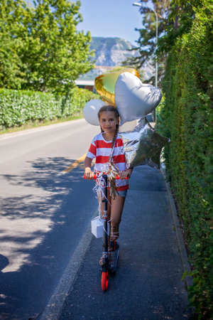 A joyful girl celebrates her birthday on a scooter in a rural setting. Colorful balloons float behind her as she rides through the sunset light. A perfect mix of freedom, youth, and adventureの写真素材