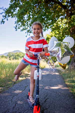 A joyful girl celebrates her birthday on a scooter in a rural setting. Colorful balloons float behind her as she rides through the sunset light. A perfect mix of freedom, youth, and adventureの写真素材