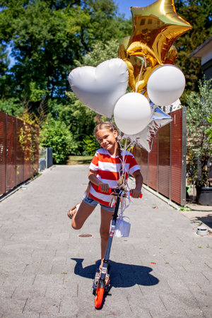 A joyful girl celebrates her birthday on a scooter. Colorful balloons float behind her as she rides through the sunset light. A perfect mix of freedom, youth, and adventureの写真素材