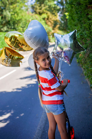 Medium shot of a cheerful girl riding her new scooter down a rustic road. Surrounded by vibrant balloons and golden sunlight, this image captures playful movement and outdoor freedomの写真素材