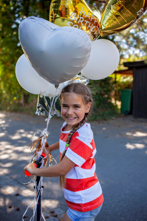 Medium shot of a cheerful girl riding her new scooter down a rustic road. Surrounded by vibrant balloons and golden sunlight, this image captures playful movement and outdoor freedomの写真素材