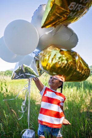 Medium shot of a cheerful girl riding her new scooter down a rustic road. Surrounded by vibrant balloons and golden sunlight, this image captures playful movement and outdoor freedomの写真素材