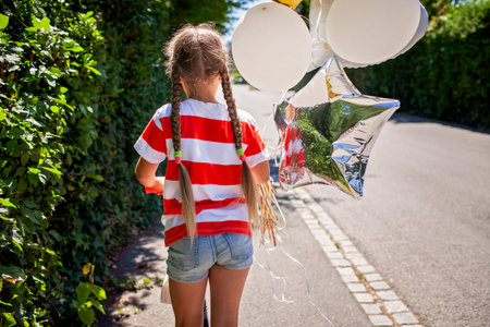 Rear view of a girl riding a scooter along a rural road at golden hour. Her birthday balloons dance in the wind. A joyful moment of carefree childhood and outdoor celebrationの写真素材