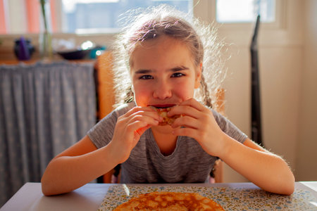 Girl happily eating a pancake with jam straight from her hands. Authentic family breakfast moment full of laughter, sweetness, and carefree childhoodの写真素材