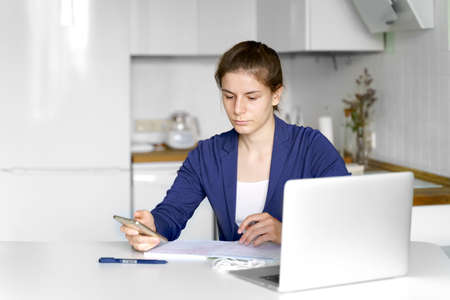 young teenage girl studying at home with laptop and phone in her hand in blue jacket