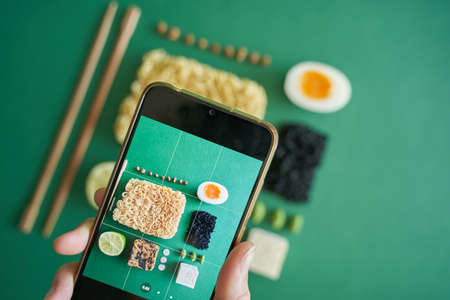 Flat lay Concept ramen soup ingredients noodle, egg, wakame algae, lime, tofu, soy meat and chopsticks and hand holding phone on green background. High quality photoの写真素材