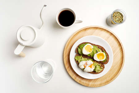 Flat lay avocado toast with eggs on dark bread, black coffee and glass of water on white background....