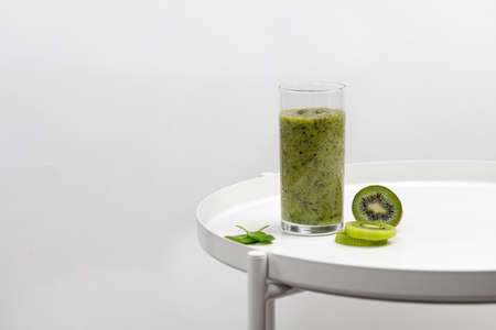 Green smoothie in a glass with slices of kiwi and spinach on white round tableの写真素材