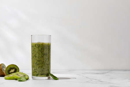 Green smoothie in a glass with slices of kiwi and spinach in the sunlight. marble table
