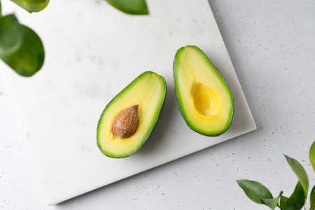 Flat lay composition with cut avocados on white marble board with green leaves. Top view, food...