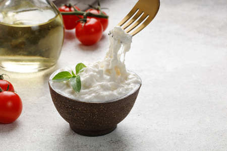 Stracciatella cheese on gray background with fork, oil, basil and tomatoes. Italian Stretched curd...