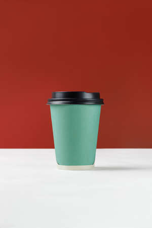 Green paper cup with coffee on white background and red wall. Disposable paper cup closeup. Blank...