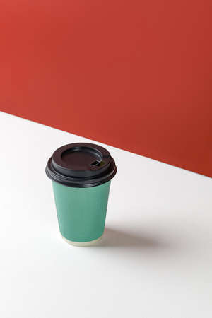 Green paper cup with coffee on white background and red wall. Disposable paper cup closeup. Blank...