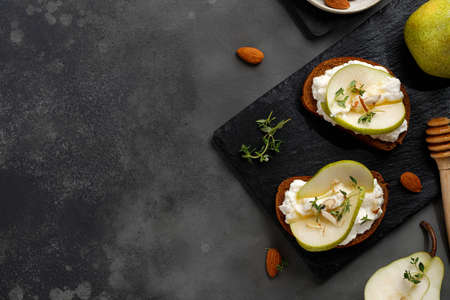 Bruschetta, snacks, appetizers. Toast with cottage cheese ricotta cheese, pear, honey and almond. Sandwiches on slate board and dark background. Copy space, text, top view.の写真素材