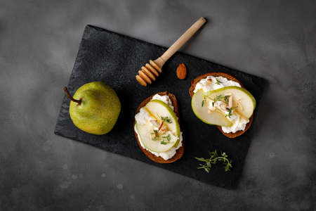 Toast with cottage cheese ricotta cheese, pear, honey and almond. Sandwiches, appetizers, bruschetta, snack on slate board and dark background. Copy space, text, top view.の写真素材