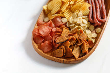 Wooden dish, plate with beer snacks, potatoes chips, meat chips, sausages kabanos, bread crackers. White background, copy space.の写真素材