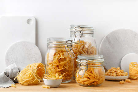 A variety of pasta in glass jars on a wooden table. White wall. Assortment raw pasta in a glass...