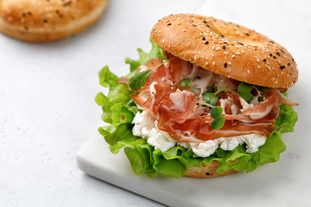 Sandwich with prosciutto, cream cheese and microgreens, bagel with ham, lettuce for breakfast on marble board. Space for text, close up.の写真素材