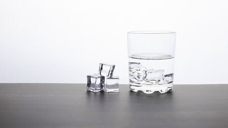 A glass with ice and water stands on a dark table against a light bright background, several ice cubes lie nearbyの写真素材