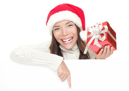 Christmas woman leaning over billboard sign. Pointing down holding gift showing big toothy smile. Caucasian / Asian woman wearing Santa hat and winter sweater isolated on white background.の写真素材