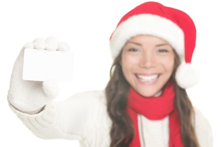Christmas woman showing business card / blank white sign with copy space. Young woman smiling in Santa hat, sweater and gloves. Isolated on white background. Mixed asian / caucasian female model. Focus on blank sign card. の写真素材