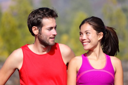 Happy sporty couple portrait. Runners outside resting after running. Man and woman smiling at each other. Asian female athlete and white caucasian male model.の写真素材