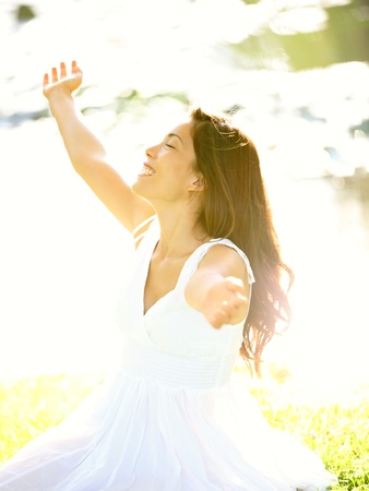 Happy free woman carefree in spring or summer with arms out enjoying the sun joyful in white summer dress sitting in the grass by lake in park. の写真素材