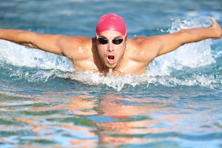 Swimmer athletic man swimming butterfly in a swimming cap approaching the camera rising up out of the water. Male athlete fitness model training outside.の写真素材